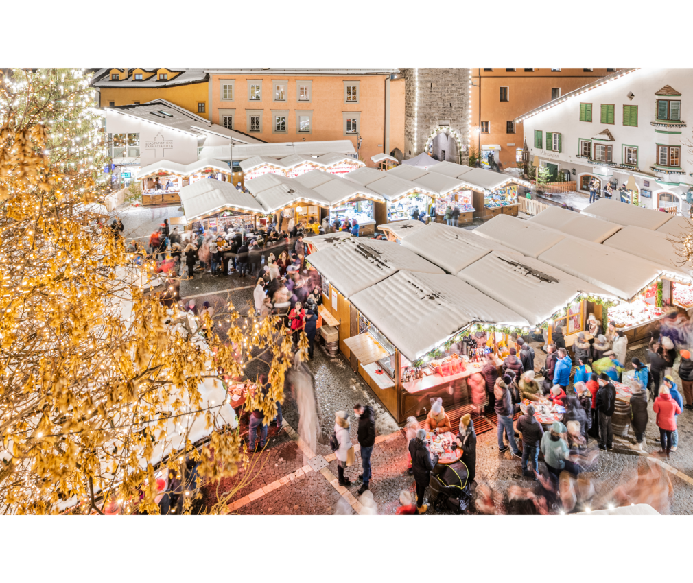 Weihnachtsmarkt Sterzing bei Tag Weihnachtsmarkt in Sterzing mit verschneiten Marktständen und regem Treiben. Im Hintergrund der beleuchtete Zwölferturm als markantes Wahrzeichen.