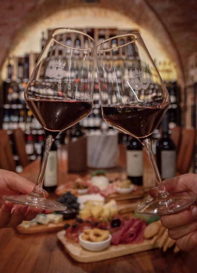 Feinkost und Wein im Mair Mair Zwei Rotweingläser im Weinkeller der Enothek Mair Mair mit Antipasti-Platten im Hintergrund. Genussvolle Weinverkostung und Kulinarik in Sterzing.