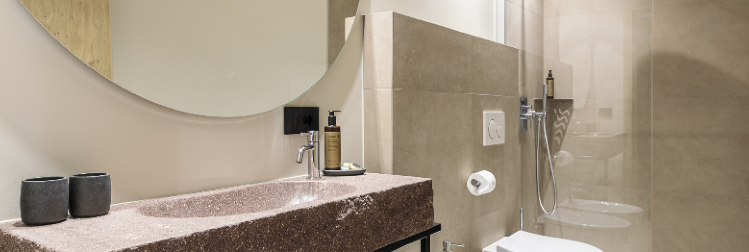 Badezimmer Porphyr Hotel Sole Modernes Badezimmer im Boutique Hotel Sole mit Porphyr-Waschbecken aus Südtirol, großem Rundspiegel, Bidet, WC und offener Regendusche in warmen Erdtönen.