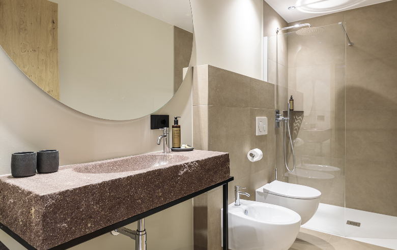 Badezimmer Porphyr Hotel Sole Modernes Badezimmer im Boutique Hotel Sole mit Porphyr-Waschbecken aus Südtirol, großem Rundspiegel, Bidet, WC und offener Regendusche in warmen Erdtönen.