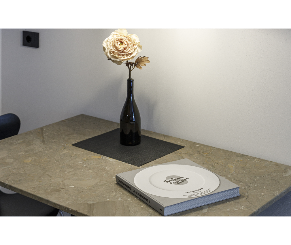 Tisch Dolomitgestein im Appartment Dolomit Minimalistisches Design im Hotel Sole: Ein edler Marmortisch mit einer schwarzen Vase und beiger Kunstblume, daneben ein stilvolles Kochbuch als Dekoration.