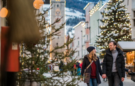 Sterzing Stadtzentrum im Winter Ein Paar spaziert Hand in Hand durch die weihnachtlich geschmückte Altstadt von Sterzing, umgeben von Lichtern, Tannenbäumen und dem historischen Zwölferturm.