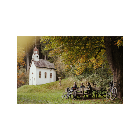 Radtour im Sterzingerumland Radfahrer rasten an einem Holztisch unter einem großen Baum nahe einer kleinen weißen Kapelle im Wald. Herbstliche Stimmung mit buntem Laub.