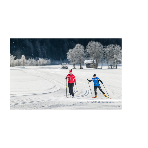 Langlaufen in Pfitsch Zwei Langläufer in sportlicher Winterkleidung auf einer gespurten Loipe in einer verschneiten Landschaft mit Bäumen und einer kleinen Hütte im Hintergrund.