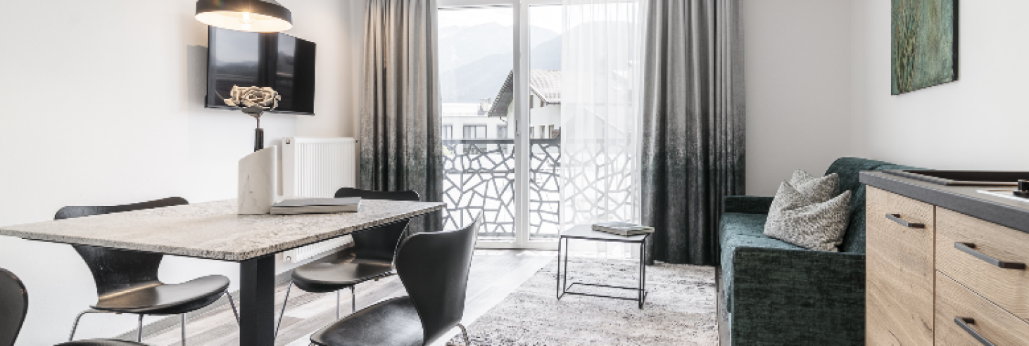 Apartment Plima Wohnraum Heller Wohnraum im Boutique Hotel Sole mit modernem Esstisch, stilvollen schwarzen Stühlen, gemütlicher Sitzecke und Balkon mit Bergblick.