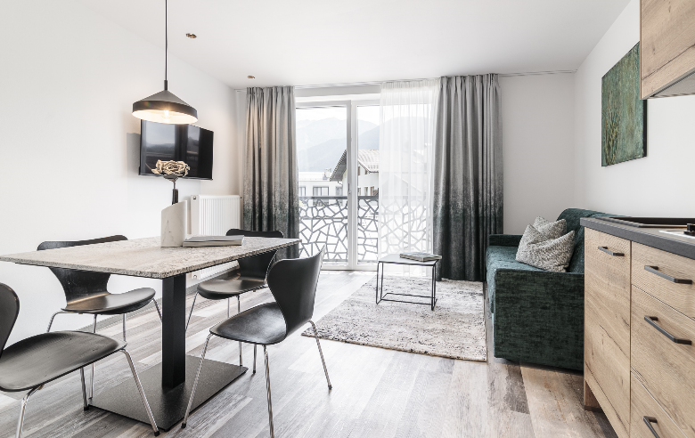 Apartment Plima Wohnraum Heller Wohnraum im Boutique Hotel Sole mit modernem Esstisch, stilvollen schwarzen Stühlen, gemütlicher Sitzecke und Balkon mit Bergblick.