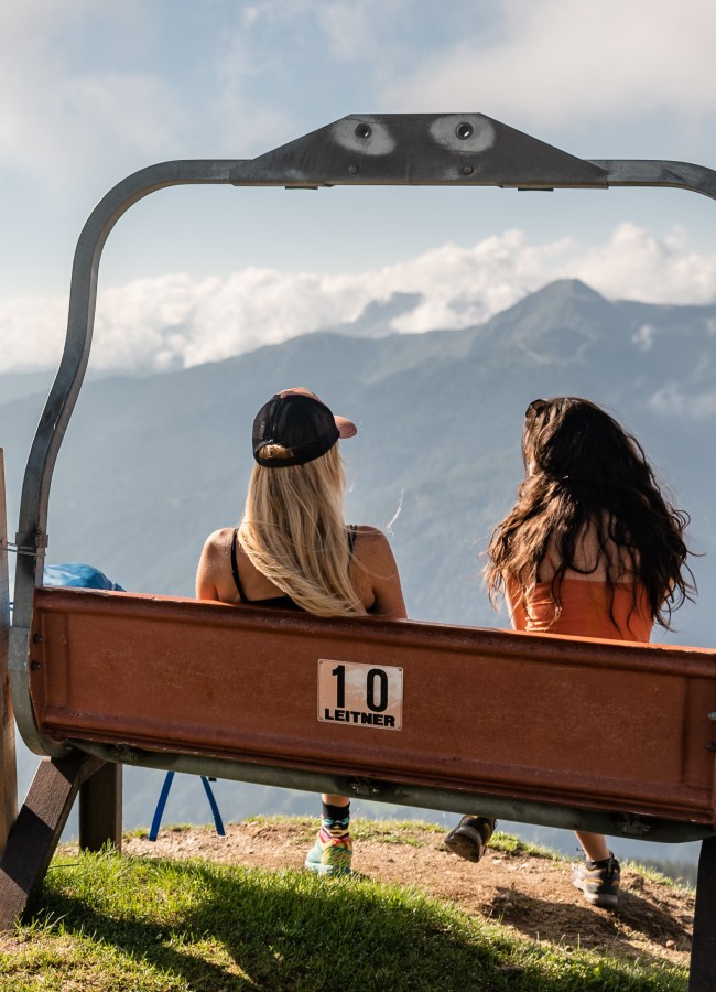 Relax am Rosskopf Zwei Wanderinnen sitzen auf einer umgebauten Sessellift-Bank der Marke Leitner und genießen den Panoramablick auf die Berglandschaft in Südtirol.