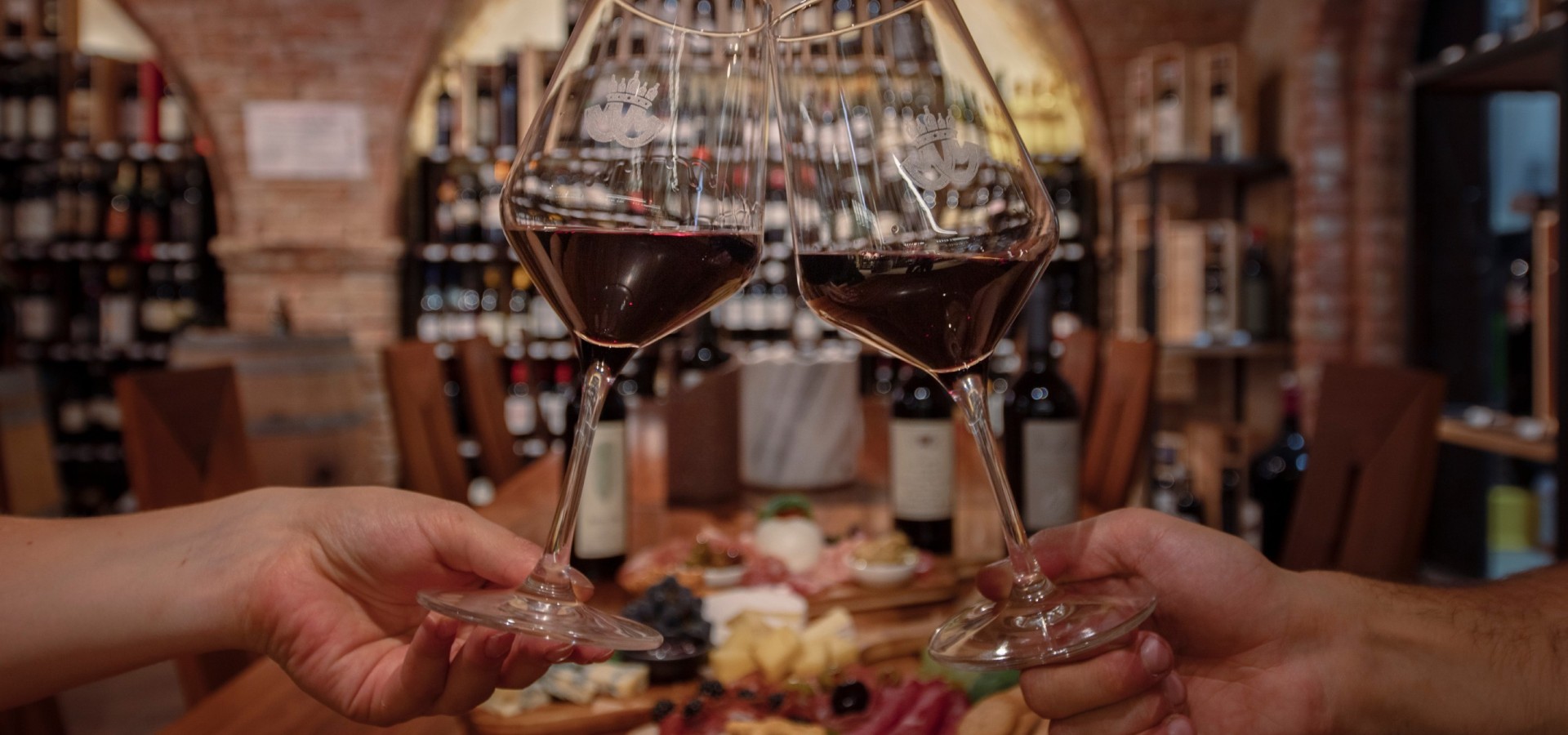 Weinverkostung im Mair Mair Zwei Personen stoßen mit Rotweingläsern, im Weinkeller von Mair Mair in Sterzing an, umgeben von Weinregalen und einer reichhaltigen Auswahl an Antipasti.