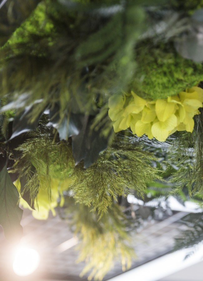 Deckenbemoosung Hotel Sole Hängende Pflanzen- und Blütendekoration in Grüntönen mit gelben Blüten. Im Hintergrund sind Lichtreflexionen und eine Deckenstruktur sichtbar.