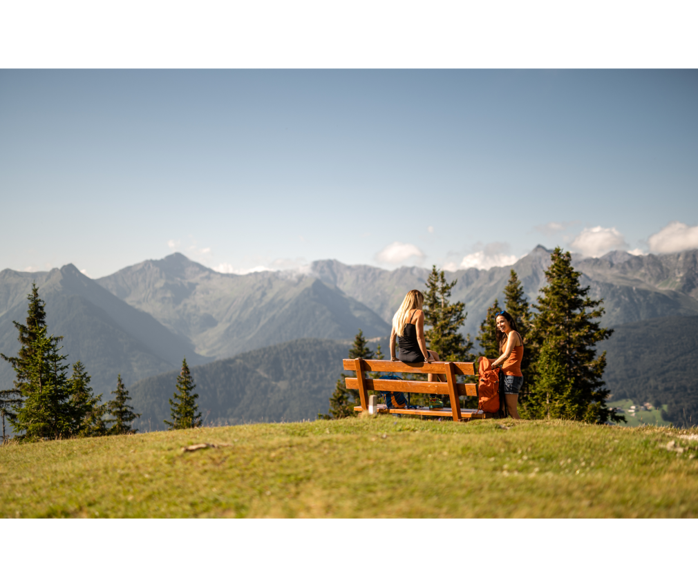 Amwiese mit Bank Zwei Frauen sitzen auf einer Bank auf einer Almwiese am Rosskopf, genießen die Aussicht auf das Ridnauntal und unterhalten sich an einem sonnigen Tag.