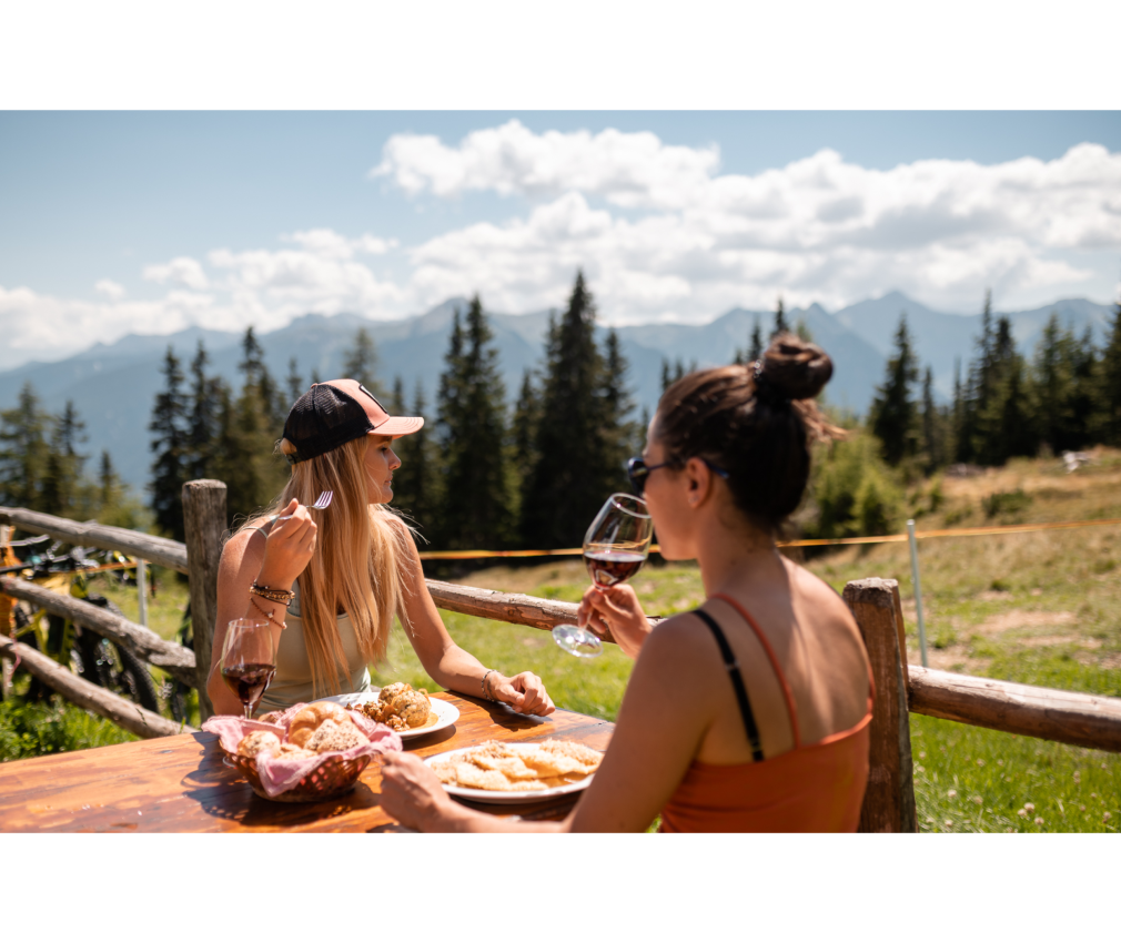 Zwei Frauen genießen eine Brotzeit und Rotwein auf einer Berghütte am Rosskopf in Sterzing mit Panoramablick auf die Südtiroler Alpen im Hintergrund.