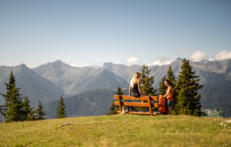 Wandern am Rosskopf Zwei Frauen genießen eine Rast auf einer Bank mit atemberaubendem Blick auf die Berglandschaft Südtirols, umgeben von grünen Wiesen und Nadelbäumen.