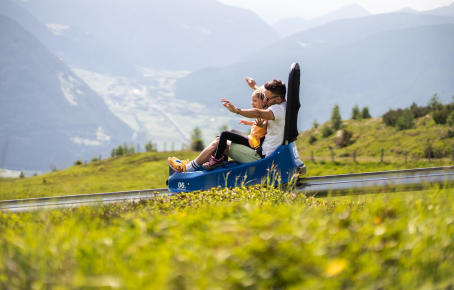 Sommerrodeln in Sterzing Vater und Tochter fahren auf einer Sommerrodelbahn durch die grüne Berglandschaft. Im Hintergrund Sterzing, das Südtiroler Wipptal und Trens.