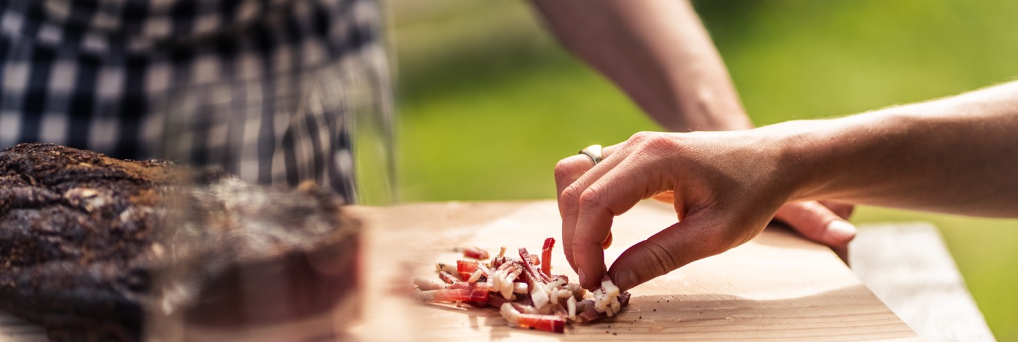 Zwei Personen bei einer gemütlichen Brettel-Jause mit geschnittenem Südtiroler Speck, einem Glas Rotwein und den Sterzinger Umland im Hintergrund